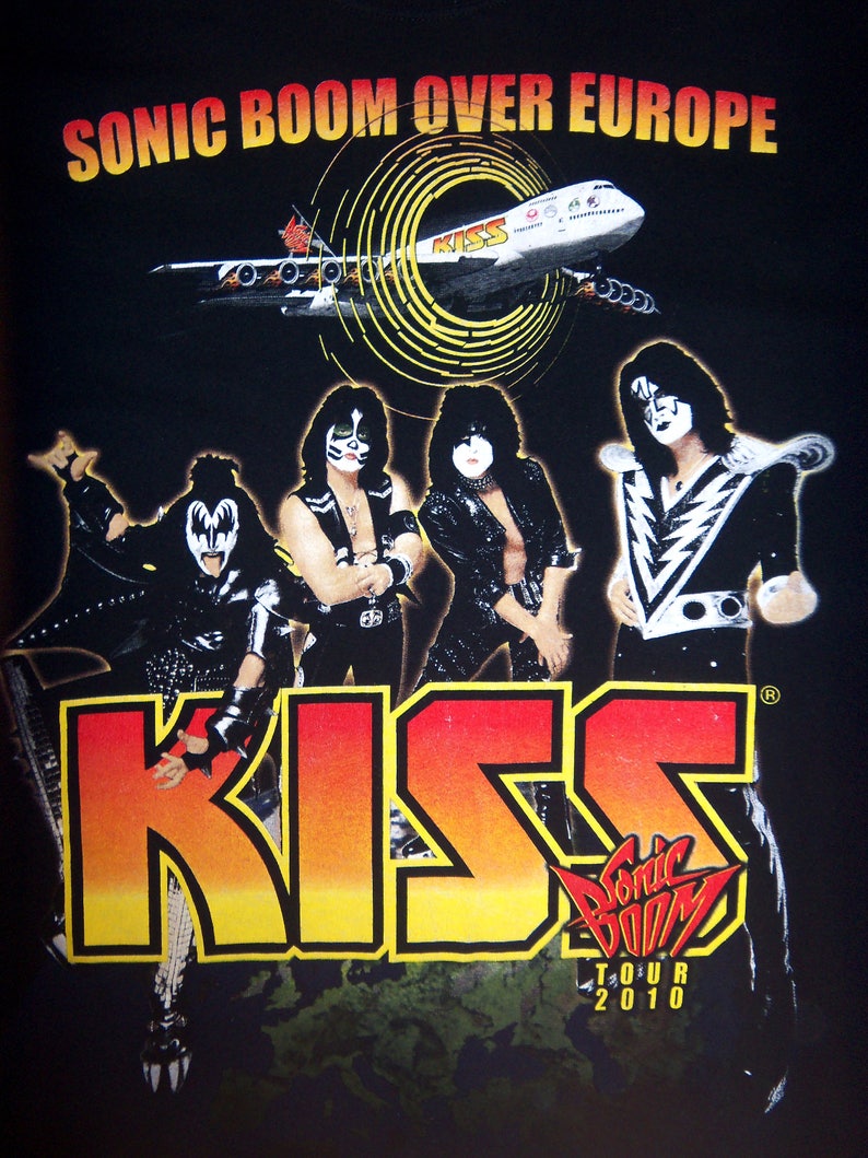 K&ouml;nnte beinhalten: Schwarzes T-Shirt mit einem Grafikdruck der Band KISS vor einem D&uuml;senflugzeug. Der Text "Sonic Boom Over Europe" befindet sich oben auf dem Shirt. Der Bandname ist in gro&szlig;en, gelben, orangen und roten Buchstaben geschrieben. Der Text "Sonic Boom Tour 2010" befindet sich unten auf dem Shirt.