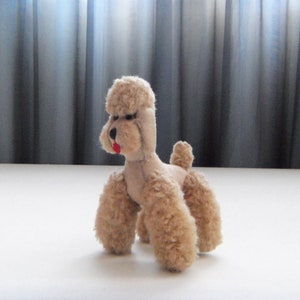May include: A plush toy poodle with a light brown fur, a pink nose, and a red tongue. The toy is sitting on a white surface.