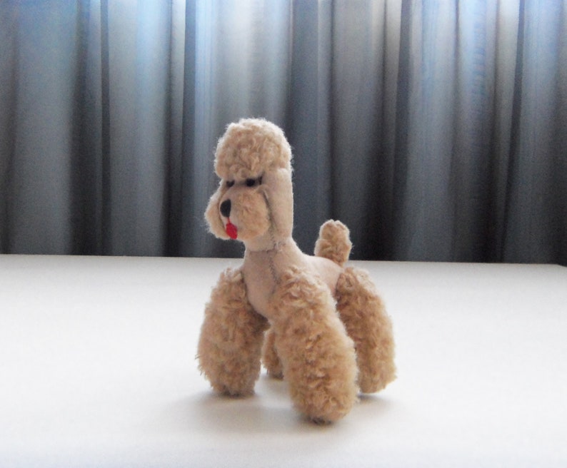 May include: A plush toy poodle with a light brown fur, a pink nose, and a red tongue. The toy is sitting on a white surface.