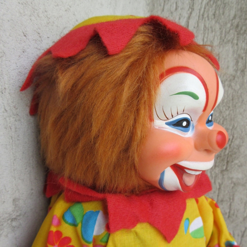 May include: A clown doll with a moulded face featuring painted white, blue, and red details, a round red nose, and a wide open-mouthed smile. The doll has textured orange hair, a red felt collar, and a yellow fabric outfit with colourful patterns.