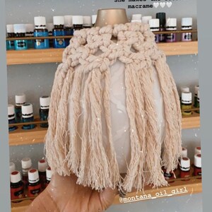 Op de afbeelding: Een witte keramische diffuser met een beige macram&eacute;-hoes. De hoes heeft een franje-ontwerp en de tekst "She makes the cutest macrame" staat boven de diffuser. De diffuser wordt in een hand gehouden.