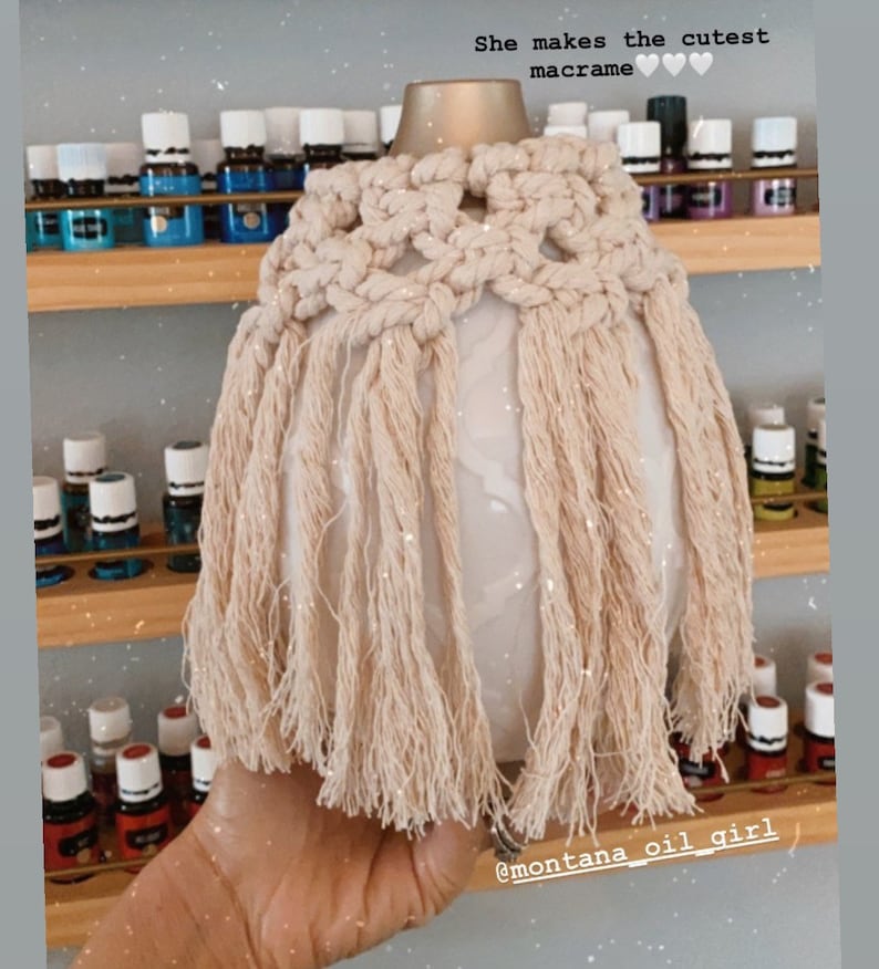 Op de afbeelding: Een witte keramische diffuser met een beige macram&eacute;-hoes. De hoes heeft een franje-ontwerp en de tekst "She makes the cutest macrame" staat boven de diffuser. De diffuser wordt in een hand gehouden.