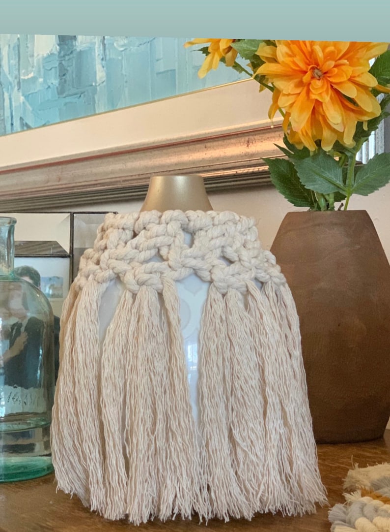 Op de afbeelding: Een witte keramische vaas met een gouden bovenkant en een cr&egrave;mekleurige macram&eacute;-bies met lange franjes. De vaas staat op een houten oppervlak.