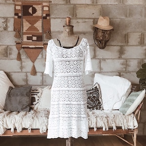 May include: White crochet dress with short sleeves and a scoop neckline. The dress is displayed on a mannequin. The dress has a geometric pattern and is knee-length. The background includes a brick wall and decorative pillows.
