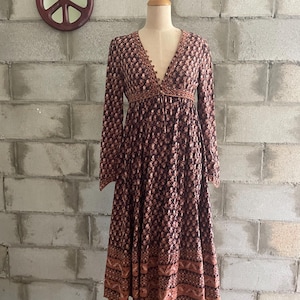 Puede incluir: Vestido de manga larga con estampado floral en tonos marrón y melocotón, exhibido en un maniquí. El vestido presenta un escote en V con detalles de botones y una falda fluida. Un símbolo de la paz de madera cuelga en la pared de ladrillo.