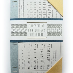May include: A small, blue and beige book with the title "Typesetting on a Winter's Afternoon" printed on the cover. The book has a decorative pattern on the front and back covers. The book is open to a page with a table of measurements and instructions for typesetting.