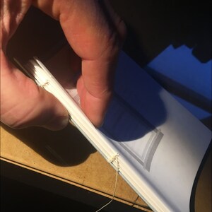May include: A white book with a blue spine is being sewn together with white thread. The book is being held by a hand.