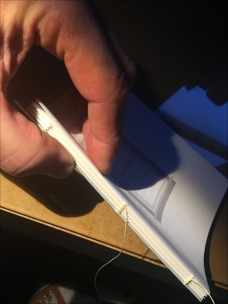 May include: A white book with a blue spine is being sewn together with white thread. The book is being held by a hand.