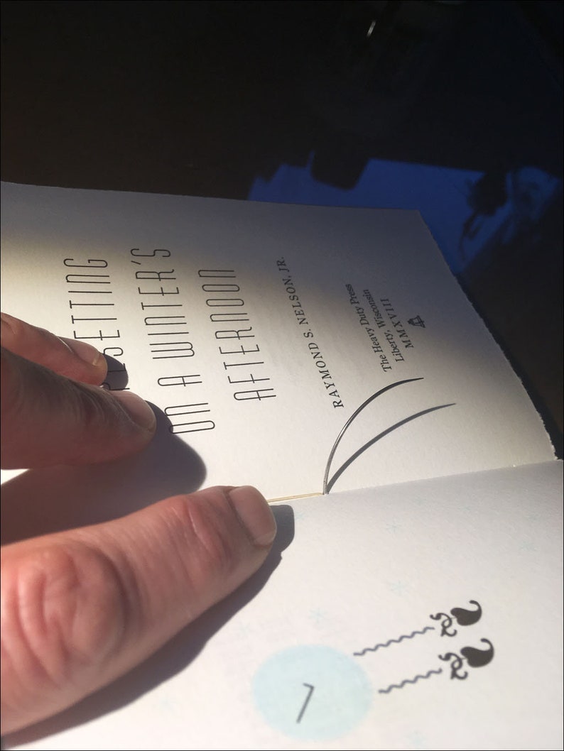 May include: A close-up of a book titled "Setting on a Winter's Afternoon" by Raymond S. Nelson, Jr. The book is open to a page with a blue circle and two black squiggly lines.