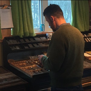 May include: A person is sorting through a drawer of metal type, which is used for printing. The drawer is part of a large, wooden printing press. The person is wearing a green sweater and is standing in a room with a window and green curtains.