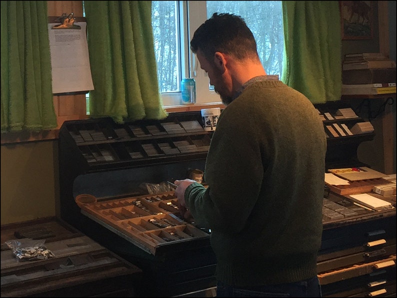 May include: A person is sorting through a drawer of metal type, which is used for printing. The drawer is part of a large, wooden printing press. The person is wearing a green sweater and is standing in a room with a window and green curtains.