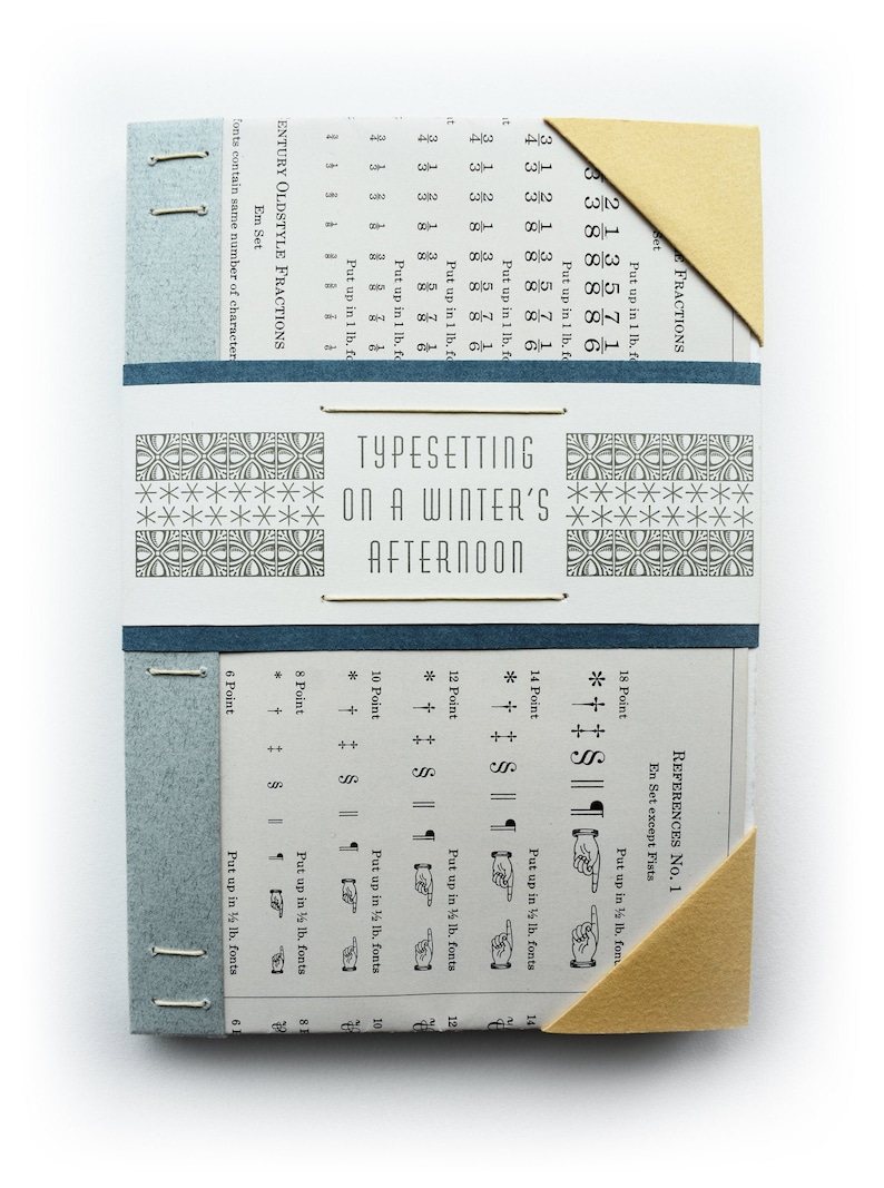 May include: A small, blue and beige book with the title "Typesetting on a Winter's Afternoon" printed on the cover. The book has a decorative pattern on the front and back covers. The book is open to a page with a table of measurements and instructions for typesetting.