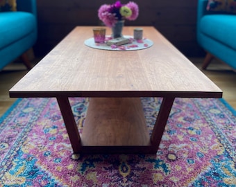 May include: A wooden coffee table with a lower shelf sits on a colorful rug in front of a window with a view of a forest. A bouquet of pink and purple flowers sits on the table.