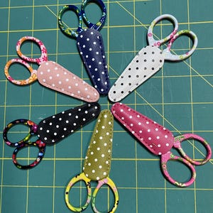 May include: A collection of small, decorative scissors with various colored handles and polka-dotted sheaths. The scissors feature floral and patterned designs on the handles and come in colors like pink, blue, black, and green. The sheaths are made of leather.