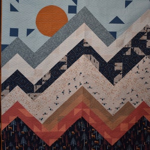 May include: A quilt with a mountain range design. The mountains are made of blue, gray, and white fabric. The background is a light blue fabric with a large orange circle in the center. The bottom of the quilt is a dark blue fabric with a pattern of trees and deer.