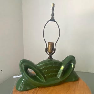 Peut inclure: Une base de lampe en céramique vert foncé présentant un design horizontal et symétrique avec des éléments latéraux incurvés en forme d'ailes et une section centrale texturée. Une douille et une armature en métal couleur laiton sont fixées.