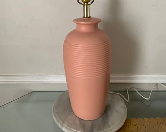 May include: A pink ceramic table lamp with a gold metal shade and a ribbed design. The lamp is sitting on a white marble surface.