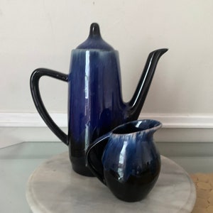 以下が含まれることがあります： 青と黒のセラミック製のティーポットとクリーマーのセット。ティーポットは背が高く、細長いボディで、側面に注ぎ口があります。クリーマーは、より小さく、丸みを帯びたポットで、ハンドルが付いています。両方のピースは光沢のある仕上げです。