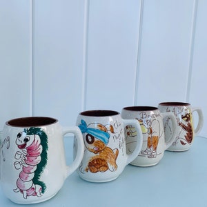 Puede incluir: Conjunto de cuatro tazas de cerámica vintage con ilustraciones caprichosas. Las tazas presentan una oruga, un perro con un parche en el ojo, una persona sosteniendo una botella y un pájaro. Las tazas son blancas con acentos marrones y rojos.