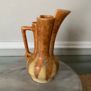 May include: A brown and yellow ceramic pitcher with a unique three-spout design. The pitcher has a textured, mottled glaze and a single handle.