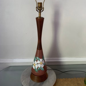 Puede incluir: Lámpara de mesa vintage con base de madera y cuerpo marrón en forma de jarrón. La lámpara presenta un diseño floral con flores blancas y hojas verdes. La lámpara mide aproximadamente 61 cm de altura.