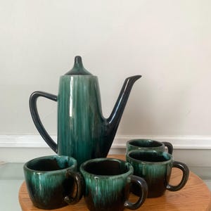 Peut inclure: Un ensemble à café vintage en céramique vert d'eau et noir. L'ensemble comprend une grande cafetière et quatre petites tasses avec des anses. L'ensemble est présenté sur un plateau rond en bois. La cafetière et les tasses ont une finition brillante.