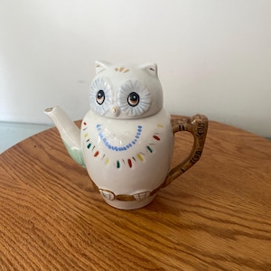May include: A white ceramic owl teapot with a decorative design. The teapot features a detailed owl face with large eyes and a lid. The body is adorned with colorful dots and a beaded necklace design. The handle is brown and the spout is white.