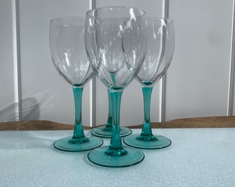 May include: Set of four clear glass wine glasses with teal green stems. The glasses are arranged in a semi-circle on a light blue surface.
