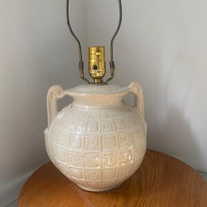 Peut inclure: Lampe de table en céramique crème avec un motif géométrique texturé. La lampe a deux petites poignées et un culot d'ampoule et un support de lampe de couleur laiton. La lampe est posée sur une table ronde en bois.