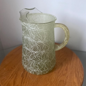 May include: A clear glass jug with a handle and a spout. The jug is decorated with a white, web-like pattern. The jug is sitting on a wooden surface.