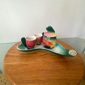 Könnte beinhalten: Ein Keramik-Servierset mit einem Erdbeerdesign in Grün, Gelb und Rosa. Das Set enthält eine Kanne, zwei Tassen und ein Tablett in Form eines Erdbeerblattes.