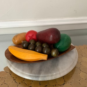 Puede incluir: Un cuenco de madera con una variedad de frutas de madera pintadas, incluyendo un plátano, uvas, una pera roja, una pera verde y un tomate rojo. El cuenco está sobre una base de mármol.