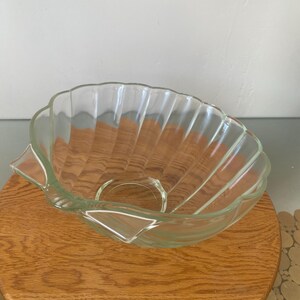May include: A clear glass bowl shaped like a seashell with a fluted design. The bowl is sitting on a wooden surface.