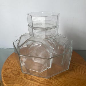 May include: A set of clear, geometric glass serving bowls. The bowls are stacked, with a large octagonal bowl at the base, followed by two smaller bowls, and a single small bowl at the top. The bowls are on a wooden surface.