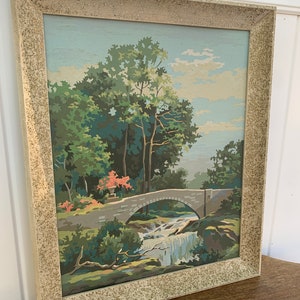 Puede incluir: Una pintura enmarcada de un puente de piedra sobre una cascada que cae en un entorno boscoso. La pintura est&aacute; realizada en un estilo pict&oacute;rico con colores brillantes y un toque de fantas&iacute;a.