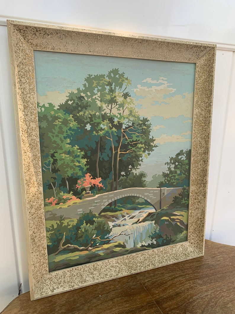 Puede incluir: Una pintura enmarcada de un puente de piedra sobre una cascada que cae en un entorno boscoso. La pintura est&aacute; realizada en un estilo pict&oacute;rico con colores brillantes y un toque de fantas&iacute;a.
