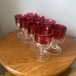 Può includere: Set di otto bicchieri da vino vintage con coppe rosso rubino e steli trasparenti e ornati. I bicchieri sono disposti su un vassoio rotondo di legno, mettendo in risalto il loro design elegante e il contrasto cromatico.