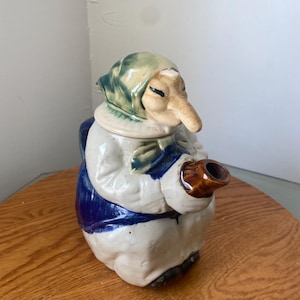 May include: A ceramic teapot in the form of a seated woman with a long nose and a green headscarf. The teapot has a white body, a blue apron, and a brown spout. The teapot is on a wooden surface.