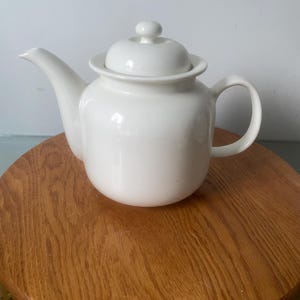 May include: A white ceramic teapot with a lid and a curved handle. The teapot is on a round wooden surface. The teapot has a spout for pouring and a small knob on top of the lid.