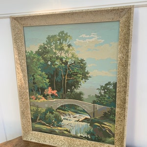 Puede incluir: Una pintura enmarcada de un paisaje con un puente de piedra sobre una cascada. La pintura est&aacute; hecha en un estilo de pintura por n&uacute;meros, con un cielo azul claro y &aacute;rboles verdes.