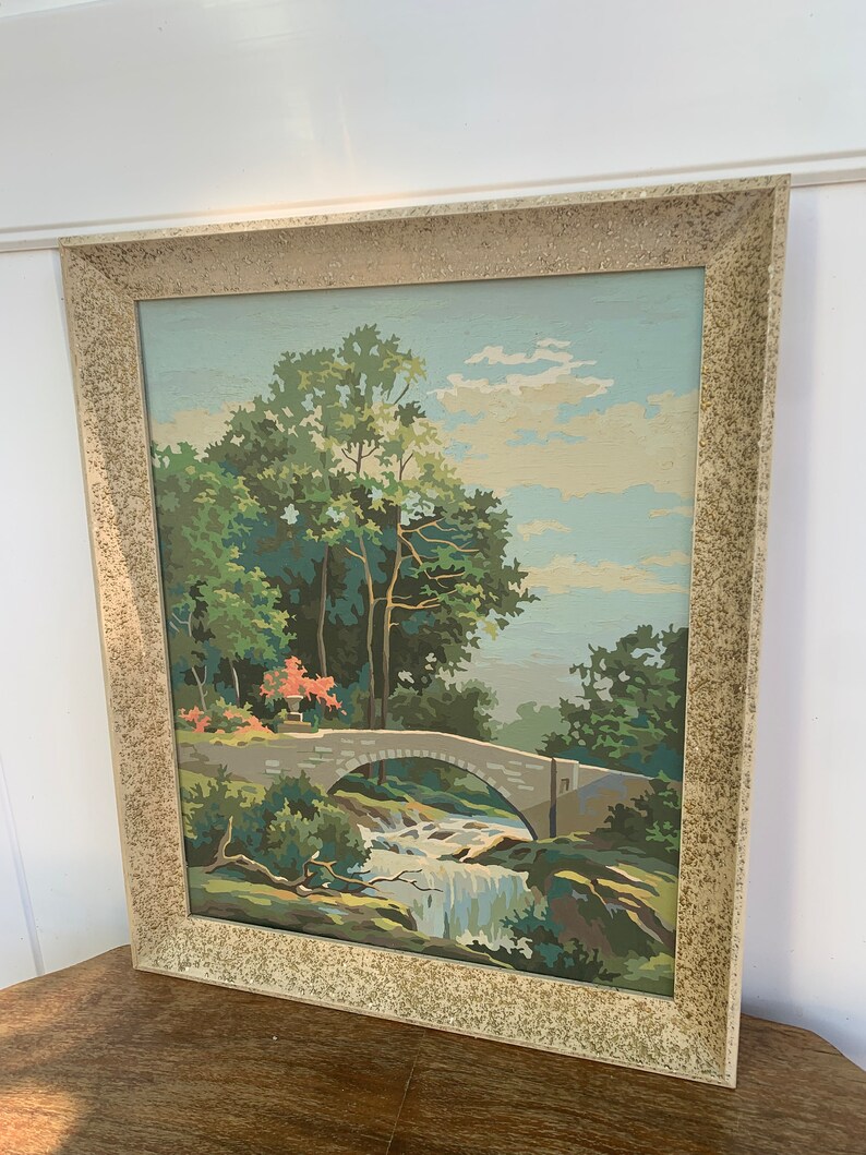 Puede incluir: Una pintura enmarcada de un paisaje con un puente de piedra sobre una cascada. La pintura est&aacute; hecha en un estilo de pintura por n&uacute;meros, con un cielo azul claro y &aacute;rboles verdes.