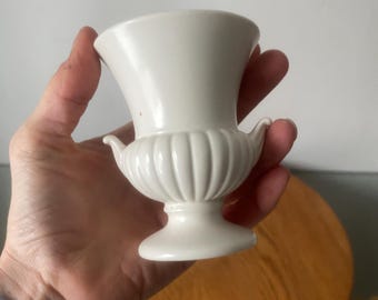 May include: A small, off-white ceramic urn with a flared top and a pedestal base. The urn features a ribbed design on the body and small, curved handles. The urn is held in a hand, with a wooden surface in the background.