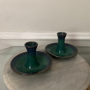 May include: Two green and blue ceramic candle holders with a small dish on the bottom. The candle holders are on a white marble surface.
