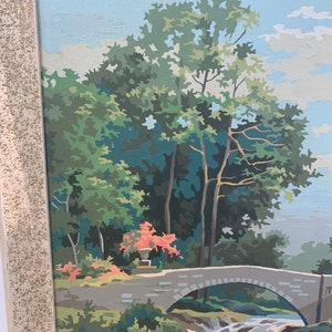 Puede incluir: Una pintura enmarcada que representa una escena de paisaje con un puente de piedra sobre una cascada. La pintura presenta un exuberante bosque verde con un peque&ntilde;o arbusto de flores rojas en primer plano. El cielo es de un azul claro con nubes blancas.