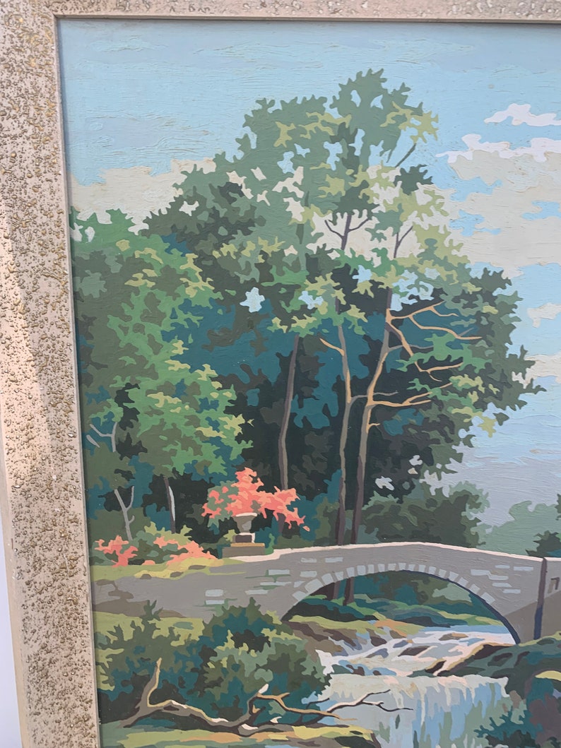 Puede incluir: Una pintura enmarcada que representa una escena de paisaje con un puente de piedra sobre una cascada. La pintura presenta un exuberante bosque verde con un peque&ntilde;o arbusto de flores rojas en primer plano. El cielo es de un azul claro con nubes blancas.