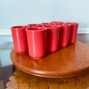 May include: A set of ten bright red ceramic shot glasses arranged in a circle on a wooden base. The glasses are all the same size and shape, and they have a smooth, glossy finish.