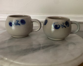 May include: Two small, round, gray stoneware mugs with blue and white painted designs. The mugs have a rounded shape and a handle on the side.