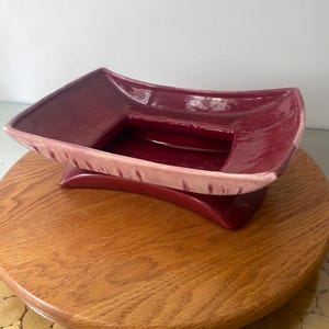 May include: A burgundy ceramic dish with a rectangular shape and a raised base. The dish has a light pink rim and sits on a wooden surface. The design is mid-century modern.