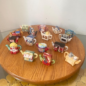 May include: A collection of fifteen miniature ceramic teapots in various shapes and colours, including designs resembling a house, a stove, a train, a person, and a shop. Each small teapot features intricate painted details and unique structural forms.