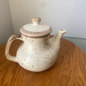 May include: A cream-coloured ceramic teapot with brown speckles, a brown handle, and a matching lid. The lid and rim have a darker brown band. The teapot is designed for brewing tea and has a spout for pouring.